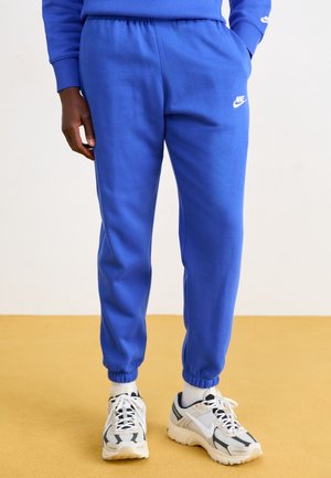 Persona vestida con pantalones deportivos Nike azul brillante, calcetines blancos y zapatillas Nike blancas y negras, de pie sobre un suelo amarillo.