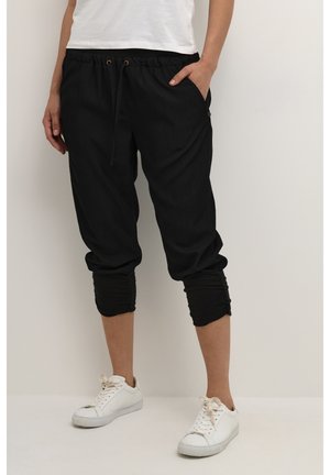 Black jogger pants made of soft fabric, featuring a cinched ankle design, elastic waistband with drawstring, and side pockets. Worn with white sneakers.