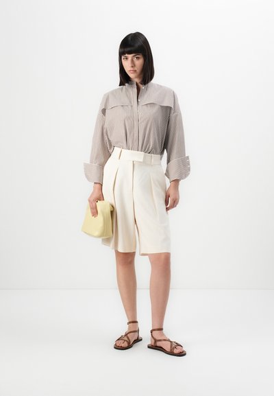 Donna con pantaloni corti plissettati beige, camicia a righe a maniche lunghe, sandali marroni, in mano una borsa giallo pallido, in piedi contro uno sfondo bianco semplice.