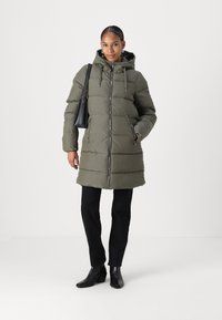 Doudoune vert olive avec fermeture éclair, capuche ajustable et poches latérales. Portée sur un pantalon noir et des bottes noires à talons.