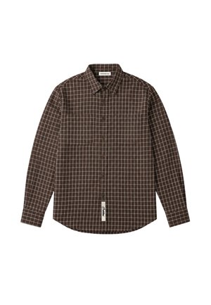 Mann trägt ein braunes kariertes Hemd mit Knöpfen, beige Hose und eine schwarze Kappe mit einem runden Aufnäher, steht mit den Händen hinter dem Rücken.