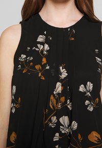 Blouse sans manches noire avec imprimés floraux blancs et marron, ornée de plis sur le devant pour apporter texture et détail au design.