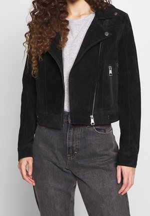 Kvinde iført en sort ruskind bikerjakke med sølv lynlåse og knapper, lysegrå skjorte og mørkegrå højtaljede jeans.