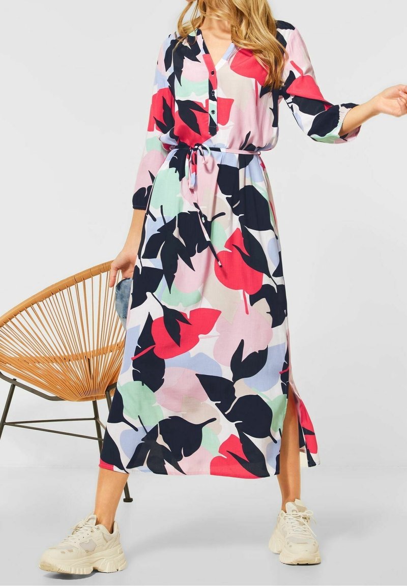Robe longue à motif feuilles multicolores avec des tons roses, verts et bleu marine, boutonnée sur le devant, ceinture à la taille et fente latérale. Portée avec des baskets.