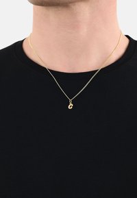 Collana in oro con un piccolo ciondolo a lettera stilizzata, indossata su una camicia nera. La superficie è liscia e lucida.