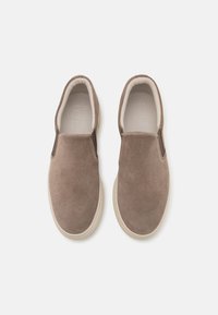 Zapatos de ante marrón tipo slip-on con acentos elásticos en los laterales, punta redonda y suela de goma blanca. Textura suave y diseño minimalista.
