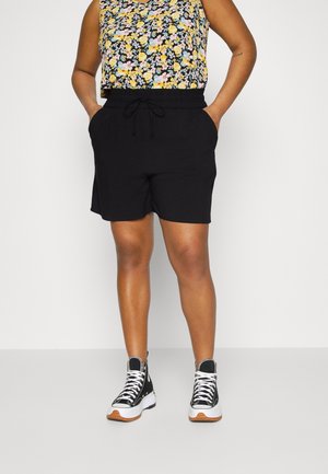 Person iført sorte shorts med snøre, blomsterprintet ærmeløs top og sorte høje sneakers med hvide såler og snørebånd, står med hænderne i lommerne.