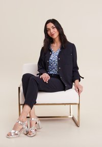 Donkerblauwe jas met knoopdetails, blouse met bloemenpatroon, zwarte broek en zilveren hoge hakken zittend op een witte stoel.