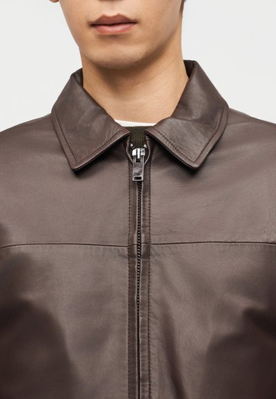 Νεαρός άνδρας που φοράει σκούρο καφέ δερμάτινο μπουφάν με φερμουάρ, εμφανίζοντας το γιακά και το πάνω μέρος του στήθους σε απλό φόντο.