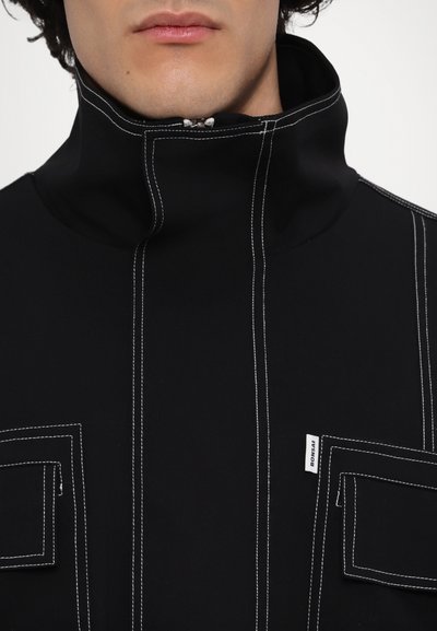 Black high-collar top with white stitching, featuring two chest pockets and a brand tag. Soft material with a smooth texture.
