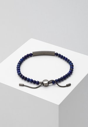 Bracelet bleu à perles avec fermoir coulissant argenté réglable et une plaque métallique rectangulaire sur un socle blanc.