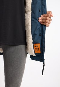 Chaqueta exterior azul con un suave forro de forro polar beige, que presenta una etiqueta naranja y botones de presión negros. También se pueden ver unos jeans grises.