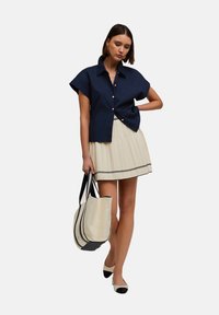 Camicia blu navy a maniche corte con bottoni dorati, gonna plissettata color crema con righe blu navy, borsa color crema con dettagli neri e scarpe bicolore.