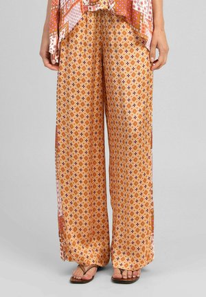 Femme portant un pantalon à jambes larges et taille haute avec un motif floral orange et des sandales marron, debout devant un fond gris uni.