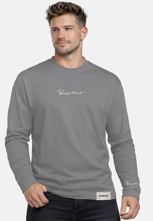 Mann trägt ein graues Langarmshirt mit weißem Text auf Brust und Handgelenk, steht mit einer Hand in der Nähe der Tasche.