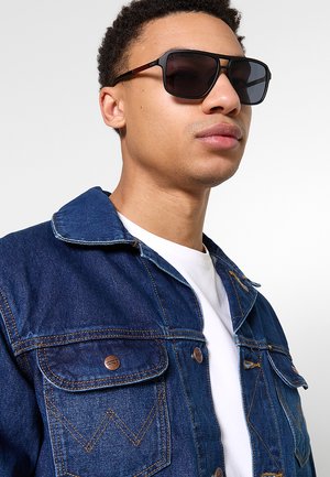 Giovane uomo con occhiali da sole neri, giacca di jeans blu con bottoni in rame e camicia bianca su uno sfondo chiaro e uniforme.
