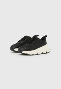 Μαύρα και άσπρα χοντρά αθλητικά παπούτσια με κορδόνια μπροστά και ανάγλυφη, χοντρή σόλα σε απλό ανοιχτό φόντο.