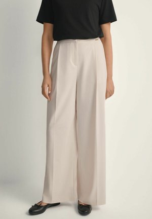 Kobieta ubrana w beżowe spodnie z wysokim stanem i szerokimi nogawkami, czarny krótki rękaw włożony do środka oraz czarne baleriny.