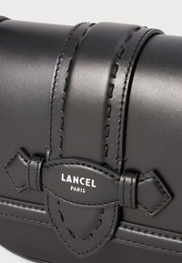 Borsa a tracolla in pelle nera con design strutturato, caratterizzata da dettagli cuciti, un accento a fiocco e etichetta logo in rilievo "LANCEL PARIS."
