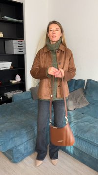 Brown suede jacket over a green turtleneck, paired with dark jeans and a brown handbag. The setting features a teal corded sofa.