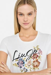 Hvid t-shirt med blomsterdesign i pink, blå og guld, med sort skråskrift "LingJo." Korte ærmer og rund halsudskæring.