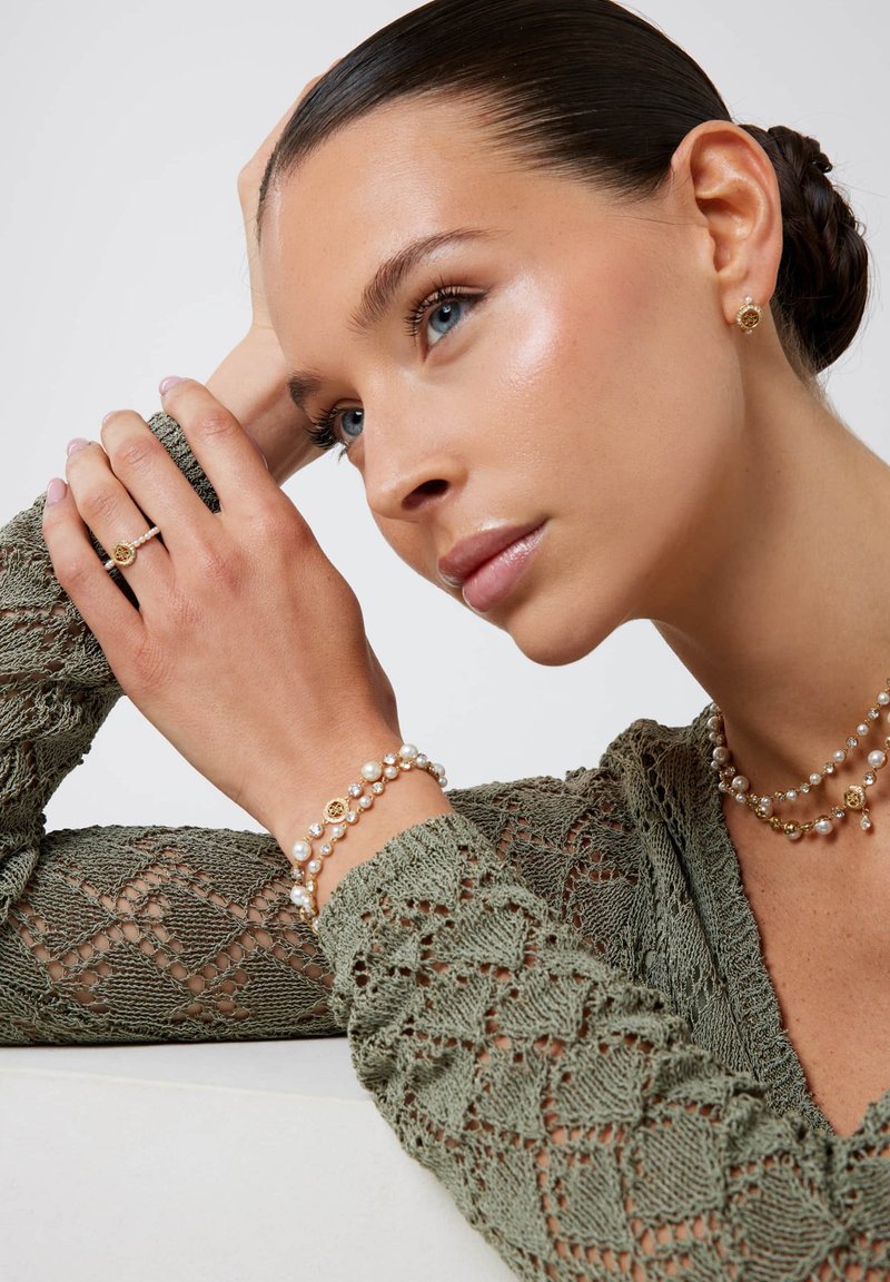 Gouden en parel sieraden op een model tonen een gelaagde ketting en armband, met een gemêleerde groene gebreide top die ingewikkelde patronen benadrukt.