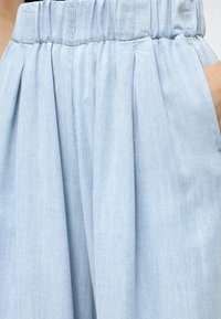 Pantalon plissé bleu clair avec une taille élastique et une main placée dans la poche droite.