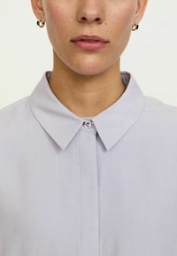 Jasnofioletowa koszula z kołnierzykiem, z zapięciem na guziki, o gładkiej strukturze i jednym guziku na kołnierzu.