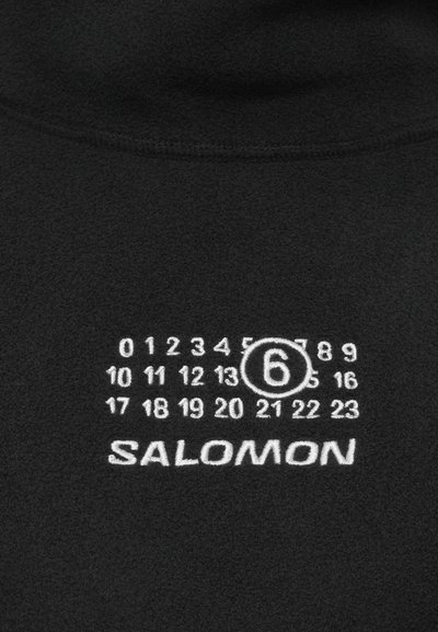 Čierna tkanina s bielymi potlačenými číslami od 0 do 23, číslo 6 je zakrúžkované, a pod číslami je značka SALOMON.