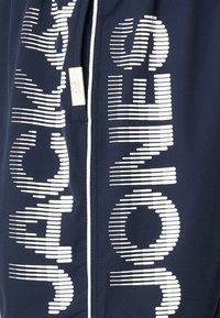 Marineblå zip-up jakke med hvit tekst og grafiske linjer. Laget av mykt, teksturert stoff med en glatt glidelås og et merke nær kragen.