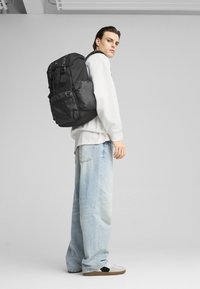 Zaino nero con multiple tasche, texture in tessuto, spallacci imbottiti, indossato da un modello maschile in una felpa bianca e jeans azzurri.