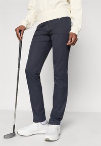 Tumed golfipüksid, mis on valmistatud kergest kangast, omavad kitsast lõiget ja peent tekstuuri. Kombineeritud valgete spordijalkadega ning hoides golfikeppi.