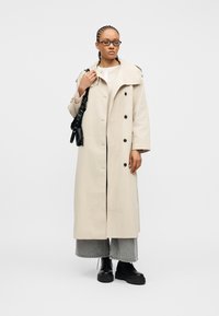 Γυναίκα με γυαλιά, ντυμένη με μακρύ μπεζ παλτό, γκρι τζιν φαρδιάς γραμμής, μαύρες μπότες, και κρατώντας μια μαύρη τσάντα ώμου, μπροστά σε απλό φόντο.