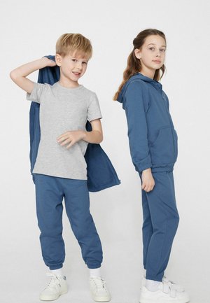 Chłopiec zdejmuje niebieską kurtkę, pod którą ma szarą koszulkę i niebieskie spodnie; dziewczynka stoi bokiem, ubrana w niebieską bluzę z kapturem i pasujące spodnie, oboje w białych trampkach.