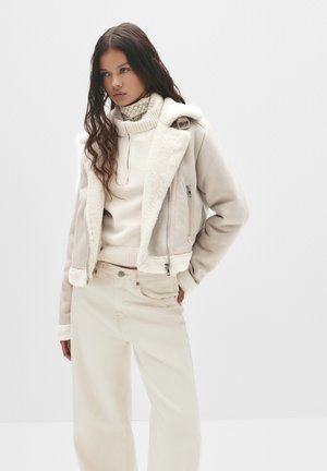 Beige Schaffelljacke mit Reißverschluss, weitem Kragen und kontrastierendem Fleece-Futter, kombiniert mit passendem cremefarbenem Pullover und weit geschnittenen Hosen.