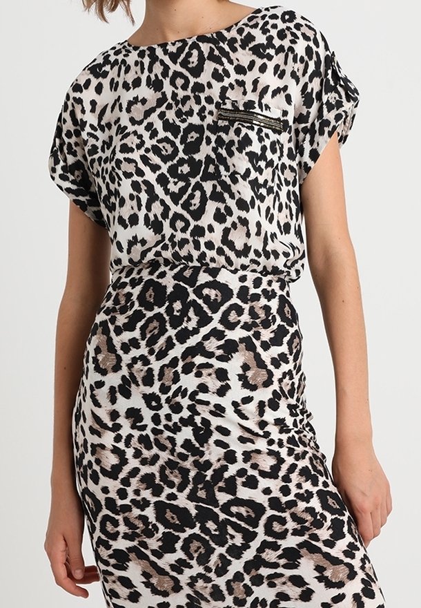 Frau trägt ein kurzärmeliges Kleid mit Leopardenmuster und einer kleinen Reißverschlusstasche an der Brust, steht vor einem schlichten hellen Hintergrund.