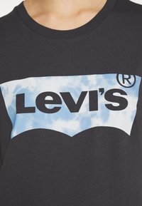 Černé bavlněné tričko s velkým logem Levi's v tučném černém písmu na pozadí modrého vzoru oblaků. Vysoký kulatý výstřih.