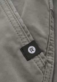 Grå stof med synlige sømme og et sort mærke med et hvidt logo, placeret ved en søm eller lomme. Blød tekstur.