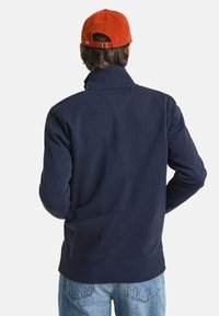 Sweat-shirt bleu marine avec un col montant, texture lisse et poignets côtelés. Associé à une casquette en velours côtelé orange, dotée d'un réglage à l'arrière.