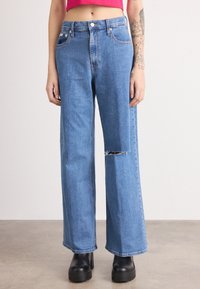 Jeans de perna larga azul claro com um joelho destru�ído, apresentando uma cintura alta, cinco bolsos e fecho de botão metálico. Combinados com botas de plataforma pretas.