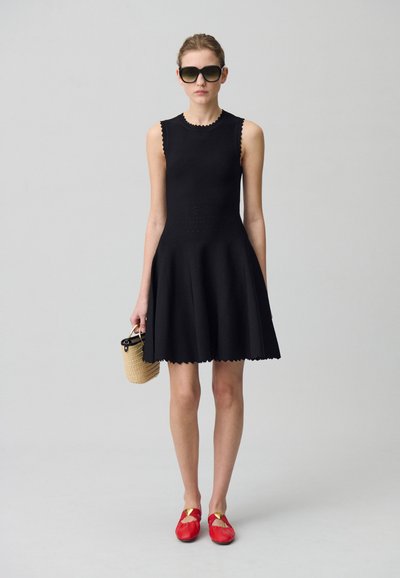 Robe noire sans manches avec une jupe évasée, un ourlet cranté et une surface texturée ; associée à des chaussures rouges et un sac en osier.