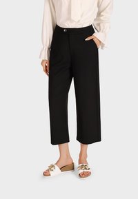 Pantaloni neri a gamba larga in tessuto liscio, con chiusura frontale a bottone e tasche laterali, abbinati a sandali bianchi con dettagli dorati.