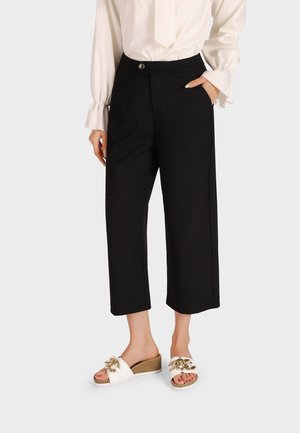 Pantaloni neri a gamba larga in tessuto liscio, con chiusura frontale a bottone e tasche laterali, abbinati a sandali bianchi con dettagli dorati.