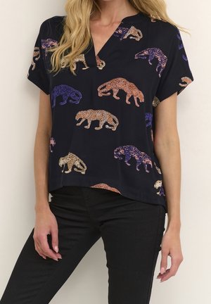 Femme portant un chemisier noir à manches courtes avec des imprimés léopard en violet, orange et beige, assorti à un pantalon noir.