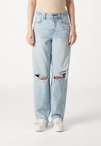 Jean en denim bleu clair à coupe ample, avec de grandes déchirures usées aux genoux et un ourlet effiloché, associés à des baskets blanches.