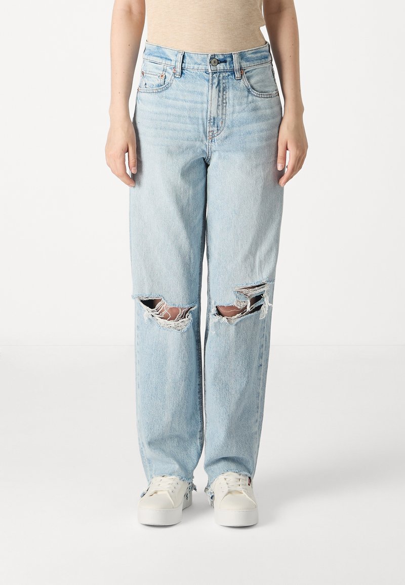 Jean en denim bleu clair à coupe ample, avec de grandes déchirures usées aux genoux et un ourlet effiloché, associés à des baskets blanches.