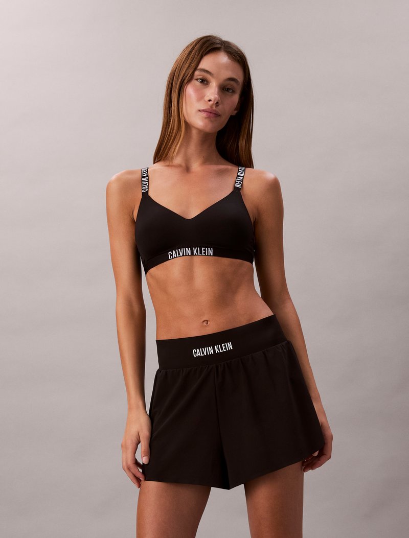 Must värvitud spordibrassi reguleeritavate rihmadega, millel on "Calvin Klein" logo, koos mustade lahtiste šortsidega elastse vöökoha ja logoga.