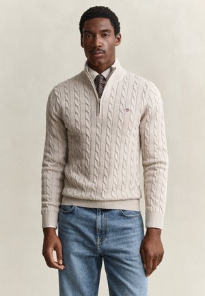 Mann trägt beigen Kabelstrick-Halbreißverschluss-Pullover über weißem Hemd und gemusterter Krawatte, kombiniert mit blauen Jeans vor einfarbigem Hintergrund.