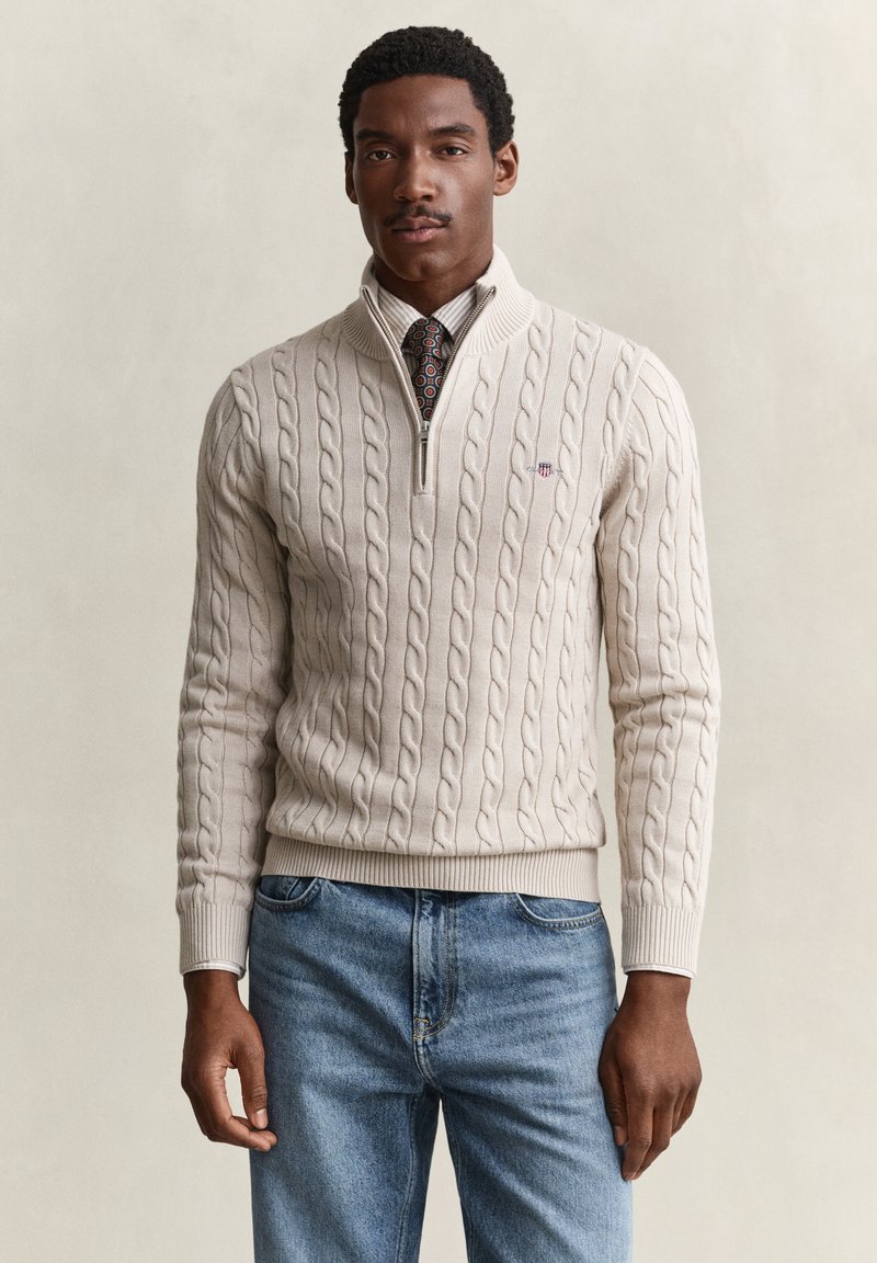 Homme portant un pull beige en maille torsadée à demi-fermeture éclair sur une chemise blanche et une cravate à motifs, associé à un jean bleu sur fond uni.