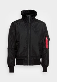 Veste bomber noire avec col en fourrure, fermeture éclair à l'avant, poignets et ceinture côtelés, poches latérales et une poche zippée avec une étiquette rouge sur la manche gauche.